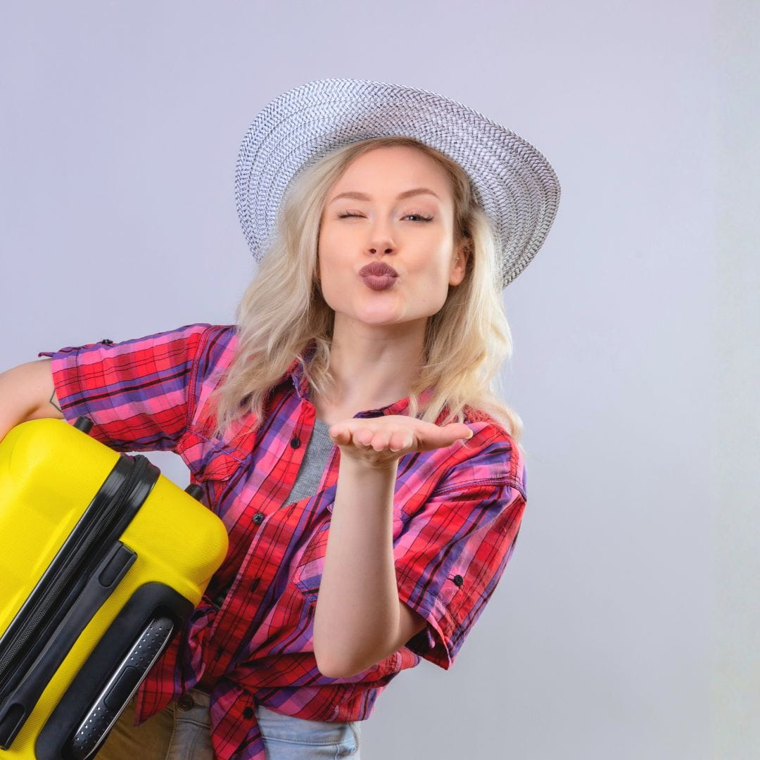 Cheerful woman blowing a kiss with one hand while holding a yellow suitcase in the other.